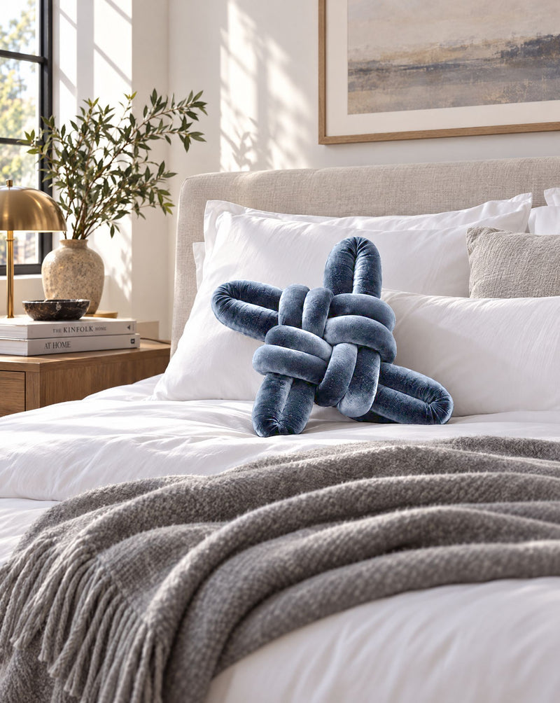 Navy blue velvet knot pillow on a white bed with a grey throw
